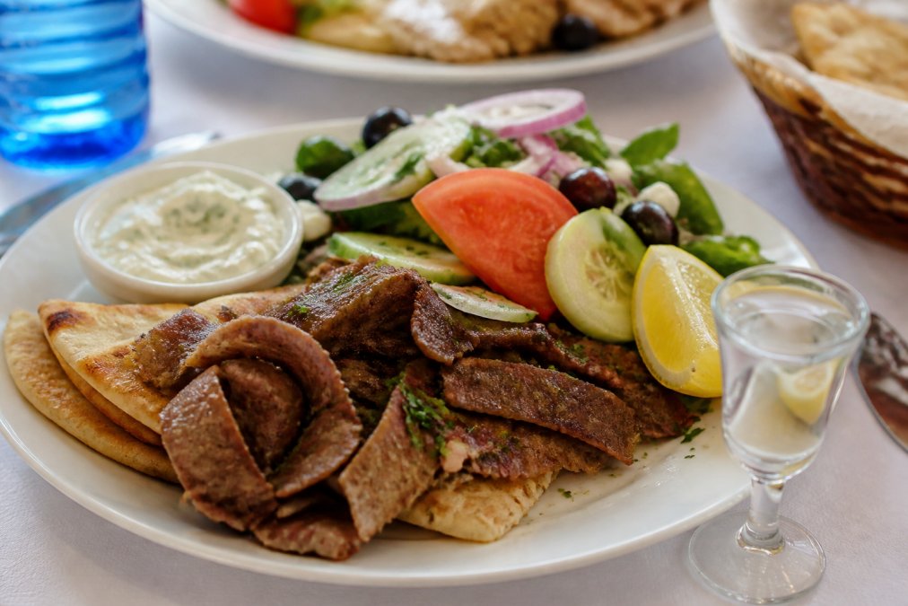 Close-up of a plate featuring traditional gyros with fresh pita bread, creamy tzatziki sauce, and colorful Mediterranean salad greens with tomatoes and cucumbers, alongside a glass of ouzo and fresh lemon wedges