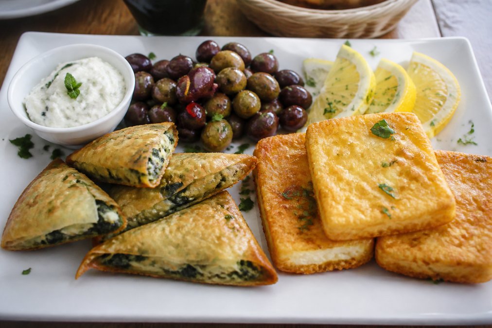 Close-up of a beautifully arranged Greek appetizer platter with golden saganaki, spinach triangles, marinated olives, fresh lemon wedges, and creamy tzatziki in a small bowl, arranged on a white ceramic plate with herbs garnish