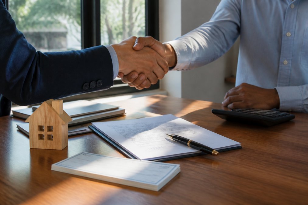 Two professionals shaking hands across desk with signed loan documents, check, and pen visible, office setting with natural light, closing moment
