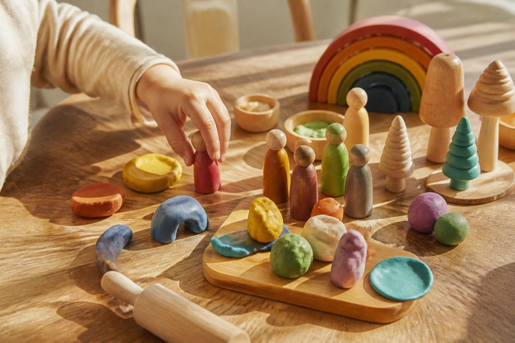 Kinderhandje speelt met natuurlijke houten speelstukken en speelklei op een houten tafel, verschillende gekleurde speelstukken en kleivormen zichtbaar, warm natuurlijk licht