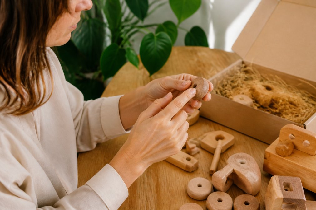 Ouder inspecteert houten speelstukken van dichtbij, voelt het hout, controleert op afwerking en splinters, keurmerklogo's zichtbaar op verpakking, warm natuurlijk licht