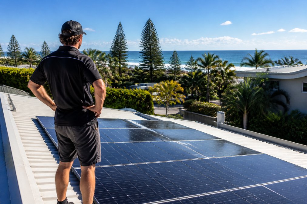 Gold Coast homeowner standing on their residential roof inspecting solar panels with ocean views and coastal palm trees visible in the background, bright midday sunlight illuminating the installation