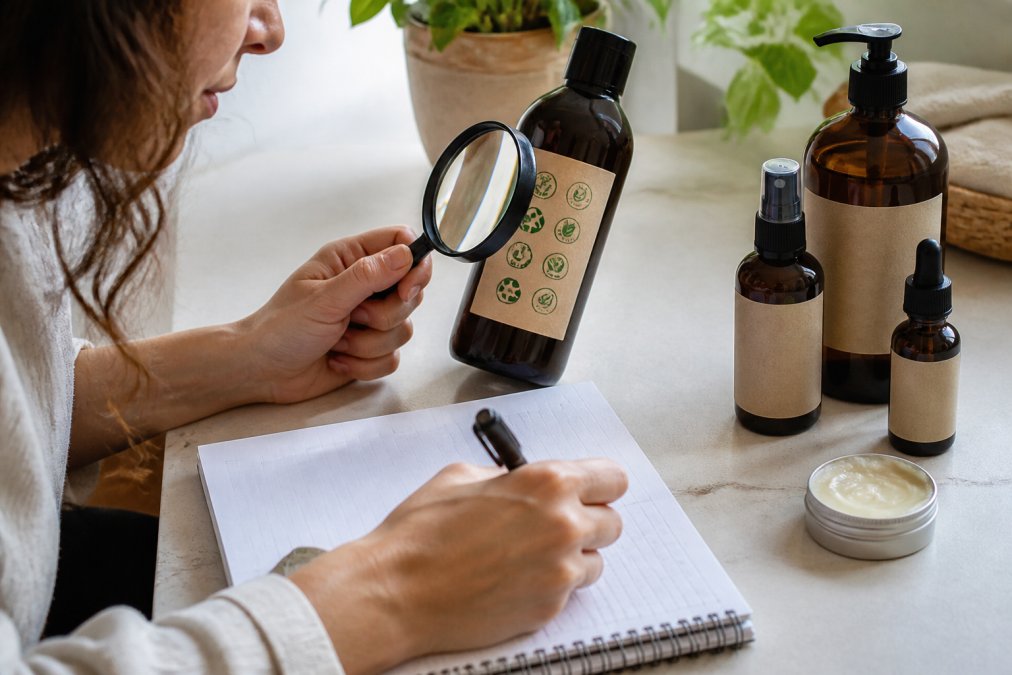 Person mit Lupe überprüft Produktverpackung mit verschiedenen Zertifikatslogos und dokumentiert Informationen mit Stift in Notizbuch auf hellem Schreibtisch