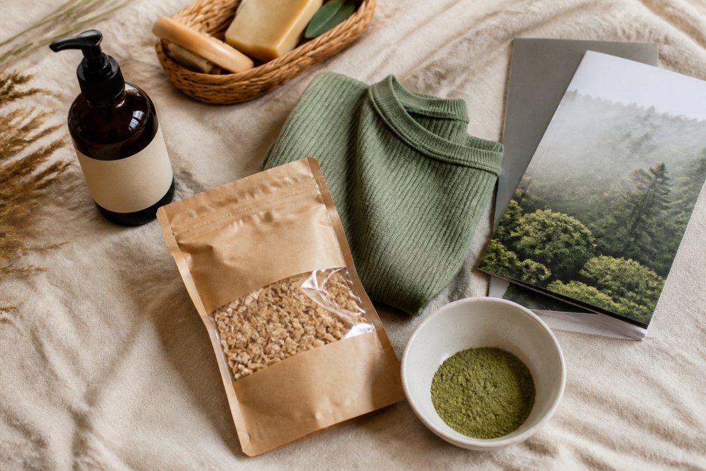 Collage aus vier verschiedenen Produktkategorien (Kosmetikflasche, Textilstoff, Lebensmittelverpackung, Finanzbroschüre), jeweils mit Greenwashing-Indikatoren markiert (vage Begriffe, fehlende Zertifikate, irreführende Prozentangaben), auf neutralem Hintergrund