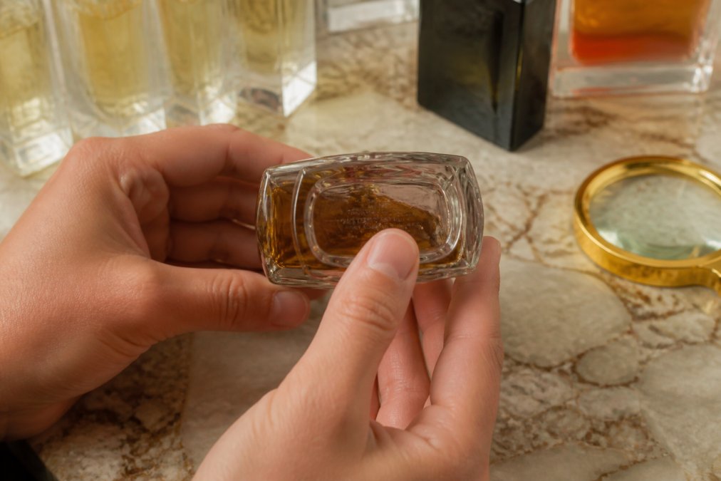 Close-up of hands examining the bottom of a perfume bottle under warm light, showing batch code and label details on a marble countertop