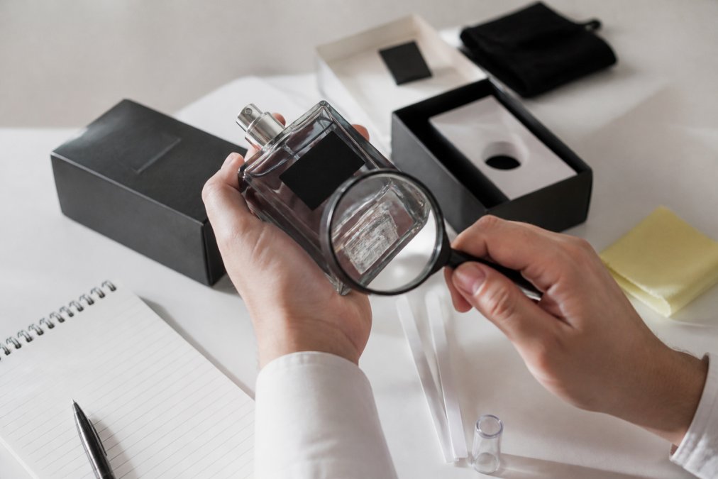 Close-up of hands carefully examining a luxury perfume bottle under bright natural light, checking the batch code label, hologram placement, and original packaging details with a magnifying glass on a white surface