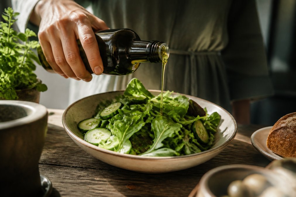 Close-up van een hand die een donkere glazen fles extra vierge olijfolie boven een heldere groene salade in een keramische kom houdt, zacht natuurlijk licht op een rustieke houten tafel