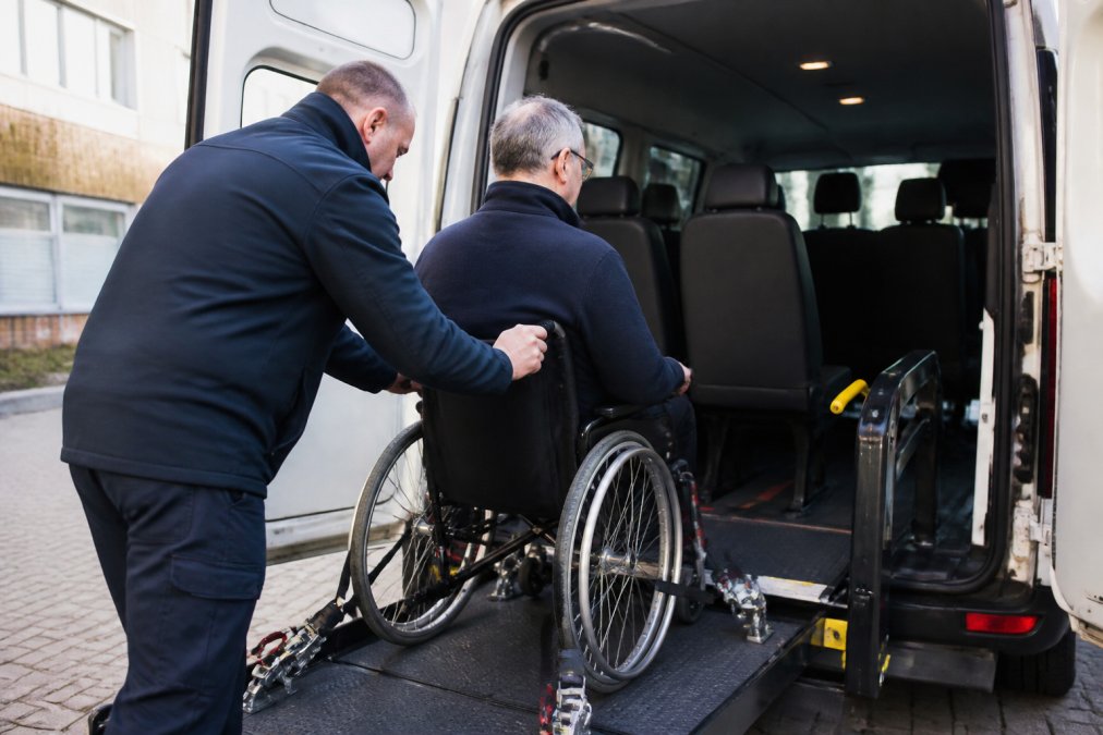Een rolstoelgebruiker wordt voorzichtig in een aangepast groepsvoertuig geholpen door een professioneel getrainde chauffeur, met zichtbare rolstoelverankering, veiligheidsriemen en comfortabele zitplaatsen in moderne klinische omgeving