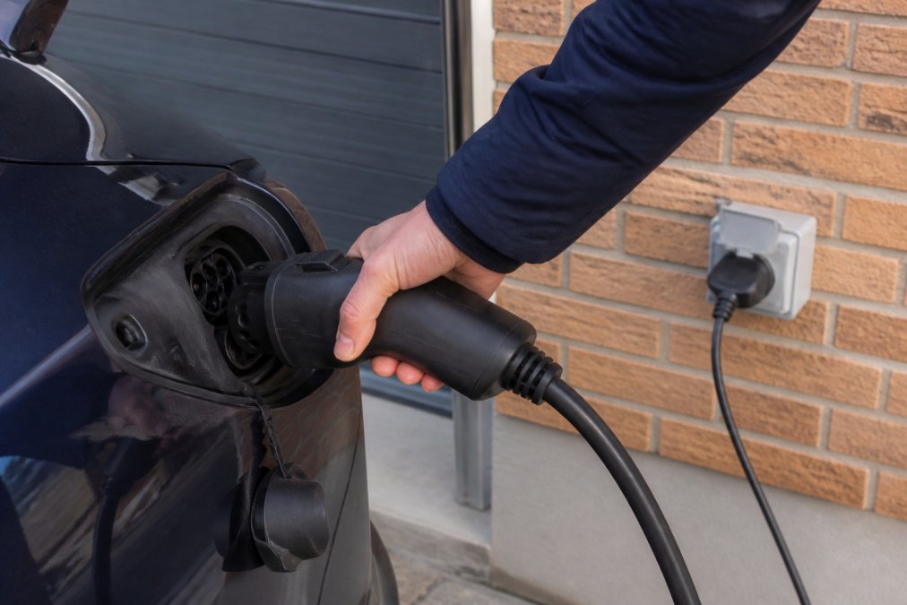 Zbliżenie adaptera V2L podłączonego do złącza portu ładowania typu 2 w samochodzie elektrycznym, w tle widoczna ściana domu i gniazdko, naturalne światło dzienne