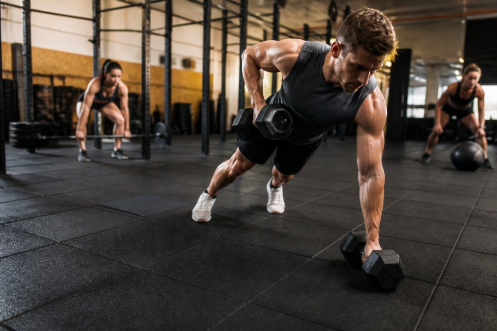 Gummigulv vs vinylgulv til træningscenter 2 Moderne fitnesscenter med sorte gummifliser på gulvet, hvor atleter løfter vægtskiver og udfører funktionel træning med intense bevægelser, tæt på en atlet der træner med håndvægte på gummigulvet
