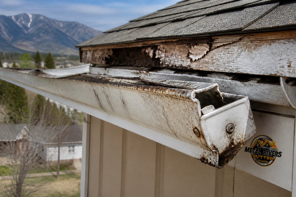 Close-up photograph of deteriorated gutter section showing rust stains, corrosion patterns, and separation from fascia board with visible water damage on the underlying wood