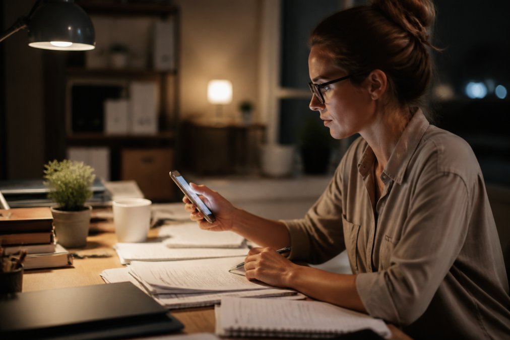 Professional sitting at desk late evening, reviewing a habit tracker on smartphone while managing multiple tasks and documents, warm desk lighting