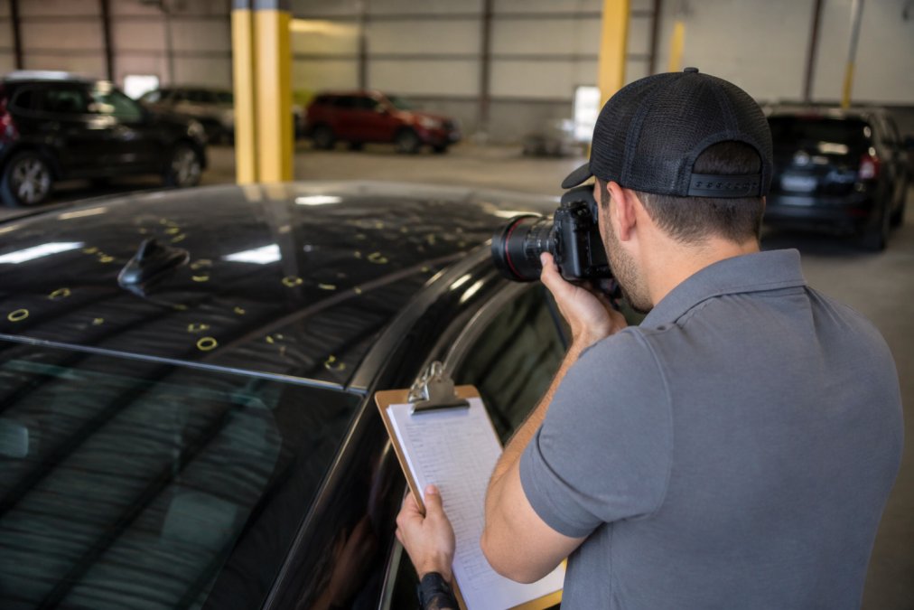Technician photographing hail damage documentation with clipboard and professional camera equipment, preparing insurance estimate in well-lit facility
