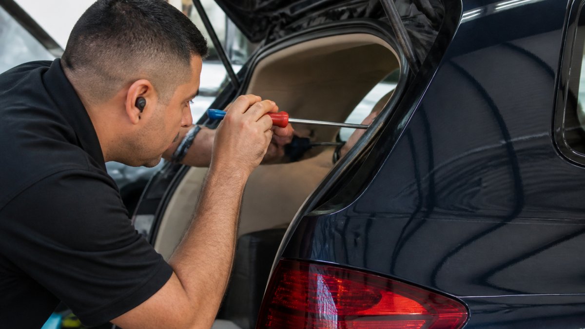 Professional technician using specialized metal rods and tools to carefully manipulate hail dent from behind vehicle panel, showing precision technique in well-lit repair facility