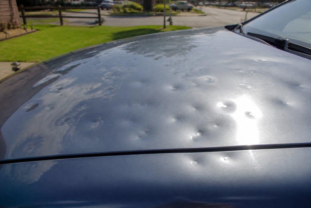 Close-up of a vehicle hood showing multiple hail dents of varying sizes, natural outdoor lighting with sunlight highlighting the impact patterns