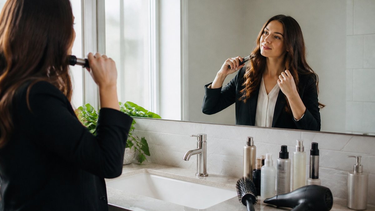 Professional woman at her bathroom mirror performing a quick morning hair styling routine, with styling tools and hair products visible on the counter, natural morning light streaming in