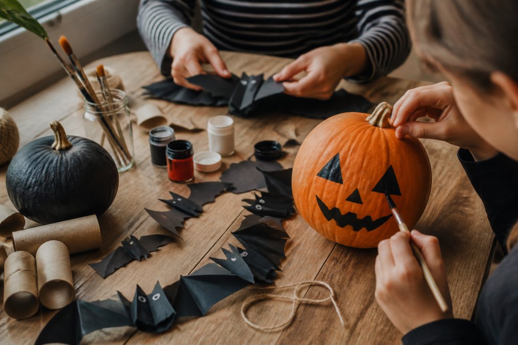 Hands crafting DIY Halloween decorations using recycled materials, folding black paper into bat shapes, painting pumpkins with acrylic paint, and arranging handmade garlands on a wooden work table