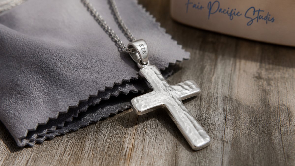 Close-up of a hammered sterling silver cross pendant resting on a weathered wooden surface next to a soft gray polishing cloth, natural window light catching the dimpled surface and a small .925 stamp near the bail