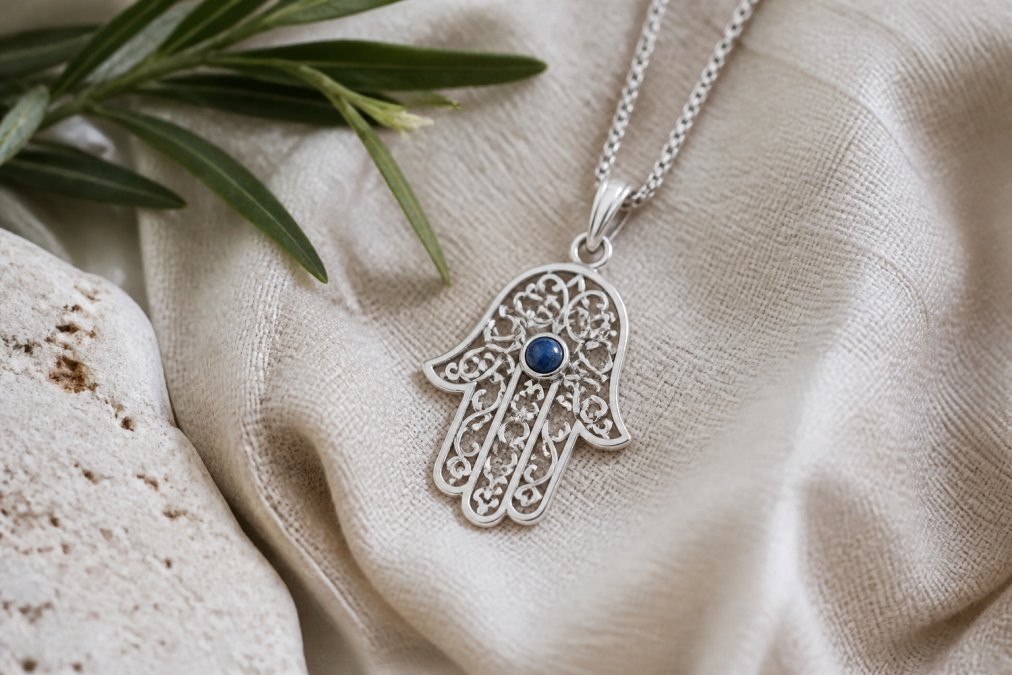 Close-up of an open sterling silver hamsa pendant with a small blue stone set in the center of the palm, resting on a linen cloth in soft daylight
