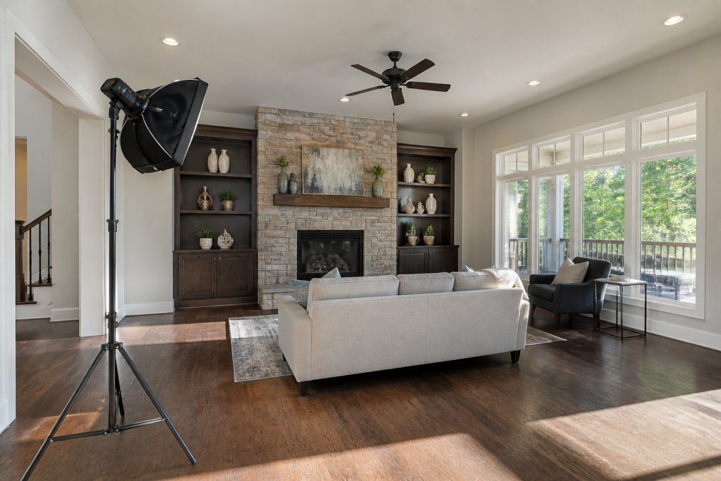Interior room setup showing professional off-camera flash positioned at 45 degrees to the left, with natural sunlight streaming through large windows on the right side, illuminating a living room with furniture and architectural details