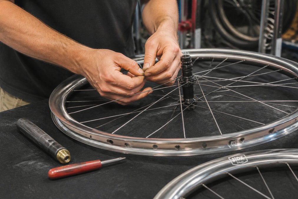 Close-up of a wheel builder's hands lacing spokes through a carbon rim on a workbench, with spoke tension tools and nipple driver visible nearby