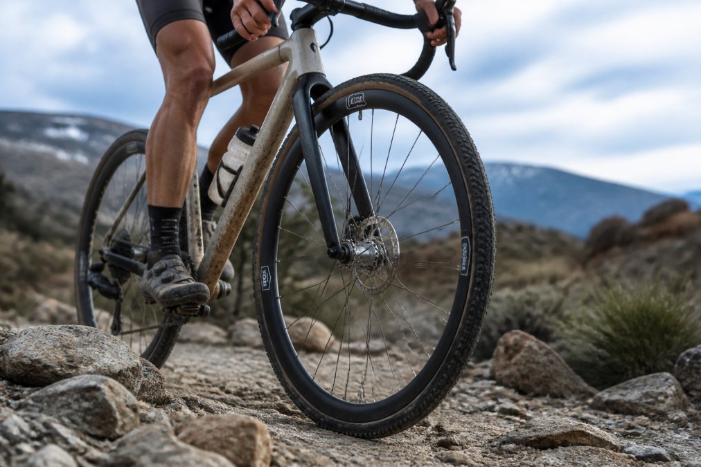 A gravel cyclist riding through rough rocky terrain, with a close-up view of a high-performance carbon wheelset showing the rim profile and polymer spokes catching the light