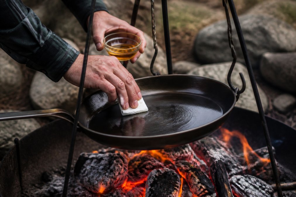 Close-up of hands applying a thin layer of oil to a carbon steel skillet suspended over glowing campfire coals, with the pan's surface catching firelight