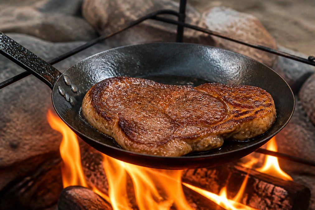 Perfectly seared steak with golden-brown crust sizzling in a carbon steel skillet suspended over bright orange campfire flames, showing even heat distribution across the meat's surface