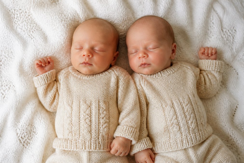 Two newborn twins lying side by side on a soft white knit blanket, each wearing a matching hand-knit cream sweater with their names embroidered in small letters at the chest, photographed from above in soft natural window light