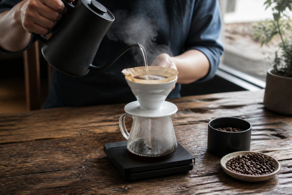Close-up of hands pouring hot water from a gooseneck kettle over a pour-over dripper, steam rising, coffee grounds blooming, rustic wooden table with a scale and coffee beans nearby