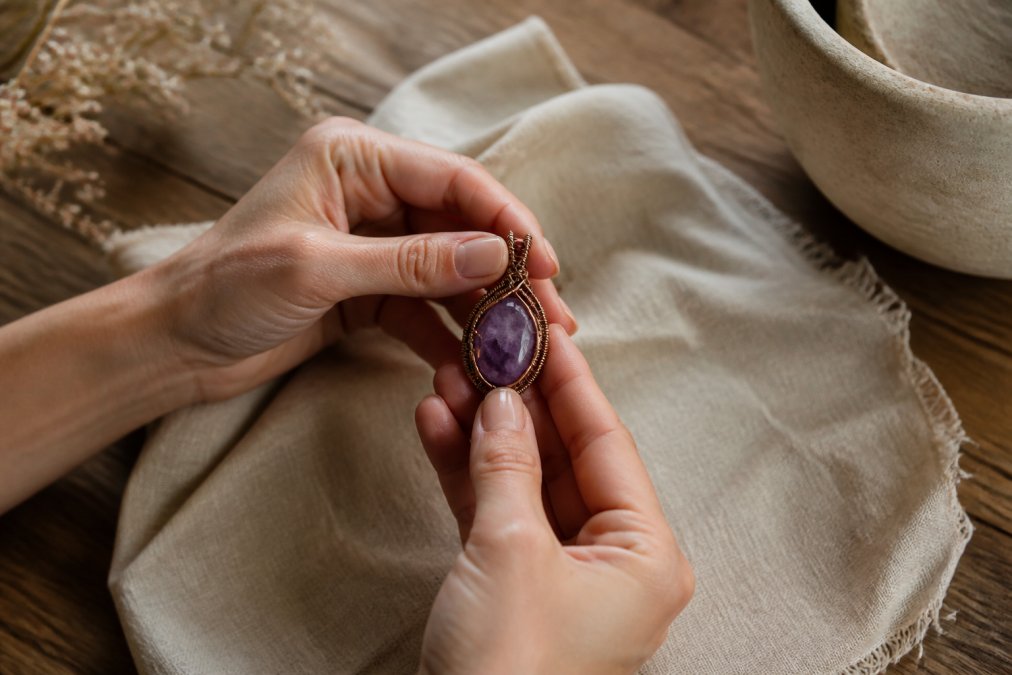 Close-up of a woman's hands holding a wire-wrapped amethyst pendant against a linen cloth on a wooden table, soft window light