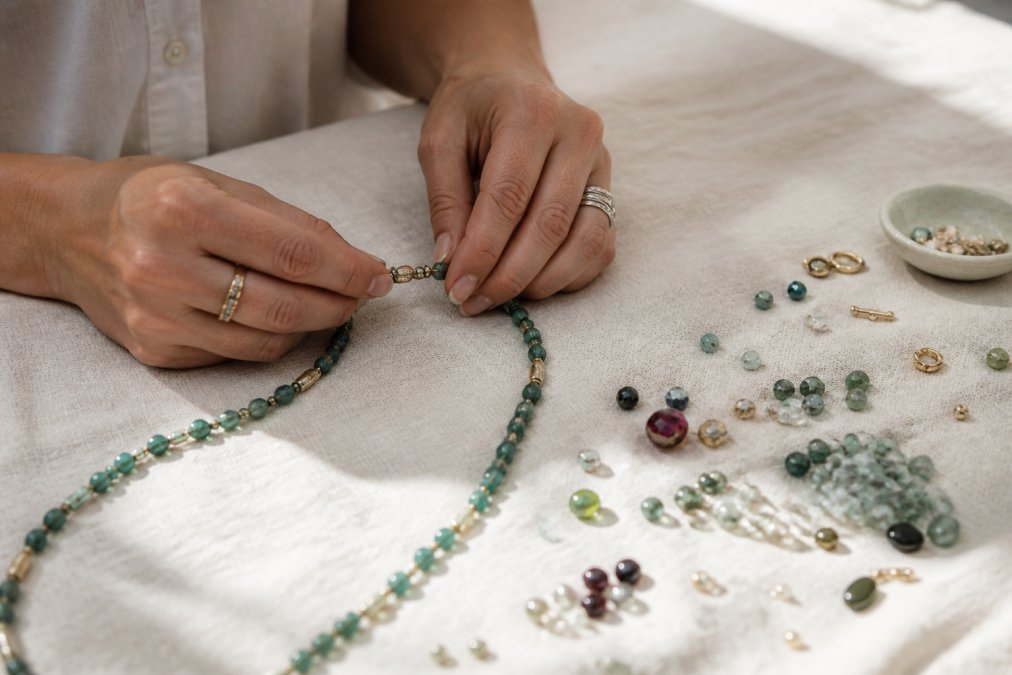Close-up of a woman's hands arranging a hand-strung gemstone necklace on a linen surface, loose gemstone beads and gold findings scattered nearby in soft natural window light