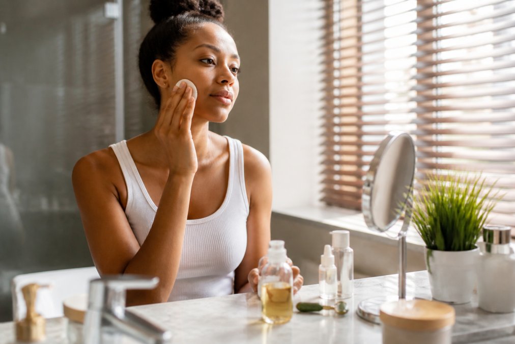 Woman applying toner to her face with a cotton pad in a bright bathroom, skincare products arranged on the vanity, warm natural light illuminating her complexion