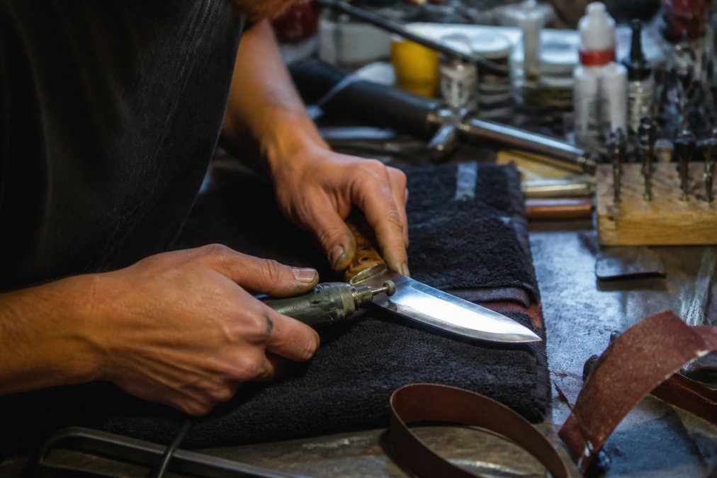 Craftsman's hands shaping and finishing a custom hunting knife blade at a workbench with specialized tools and materials visible under workshop lighting