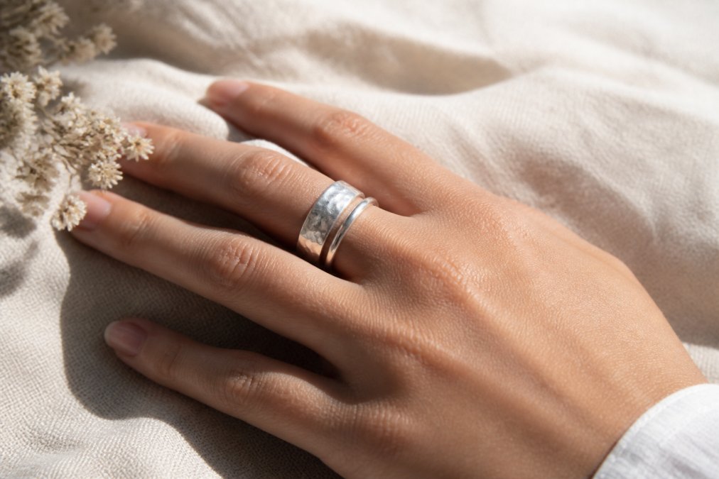 A woman's hand wearing a wide hammered sterling silver band ring and a narrow band ring stacked together, photographed against a neutral linen background with soft natural daylight coming from the side