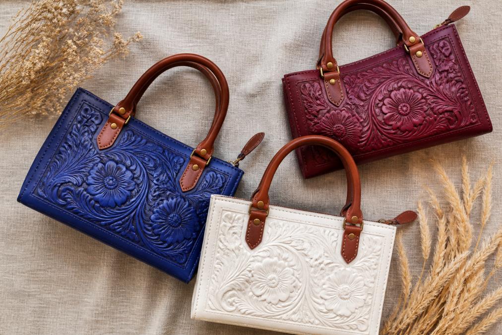 A flat lay photo of three hand-tooled leather purses in royal blue, pearl white, and burgundy on a neutral linen background, showing the detailed floral carving and cognac leather handles