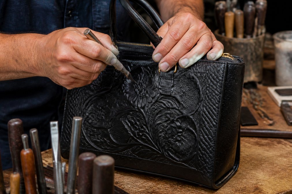 Close-up of artisan hands using traditional carving tools to create hand-chiseled floral patterns on genuine leather, showing precision detail work and the texture of hand-tooled design