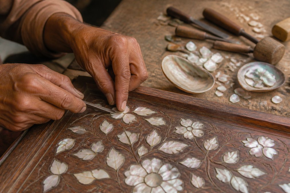 Close-up of an artisan's hands pressing a small piece of iridescent shell into a carved recess on a wooden cabinet panel, traditional hand tools and shell fragments scattered on the workbench, warm workshop lighting
