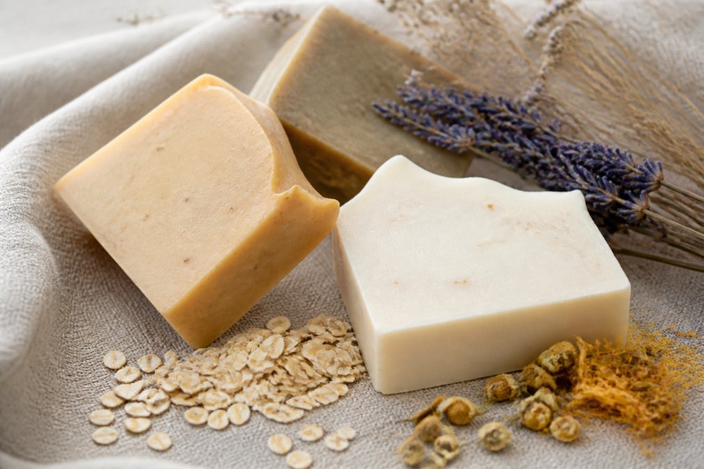 Artisanal soap bars arranged on a natural linen surface with dried botanicals and oatmeal scattered nearby, showing texture and craftsmanship in soft natural light