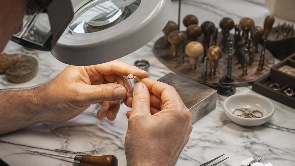 Close-up of a jeweler's hands examining a fine gold ring under bright magnifying light, inspecting the stone setting, metal edges, and polishing quality on a white marble work surface