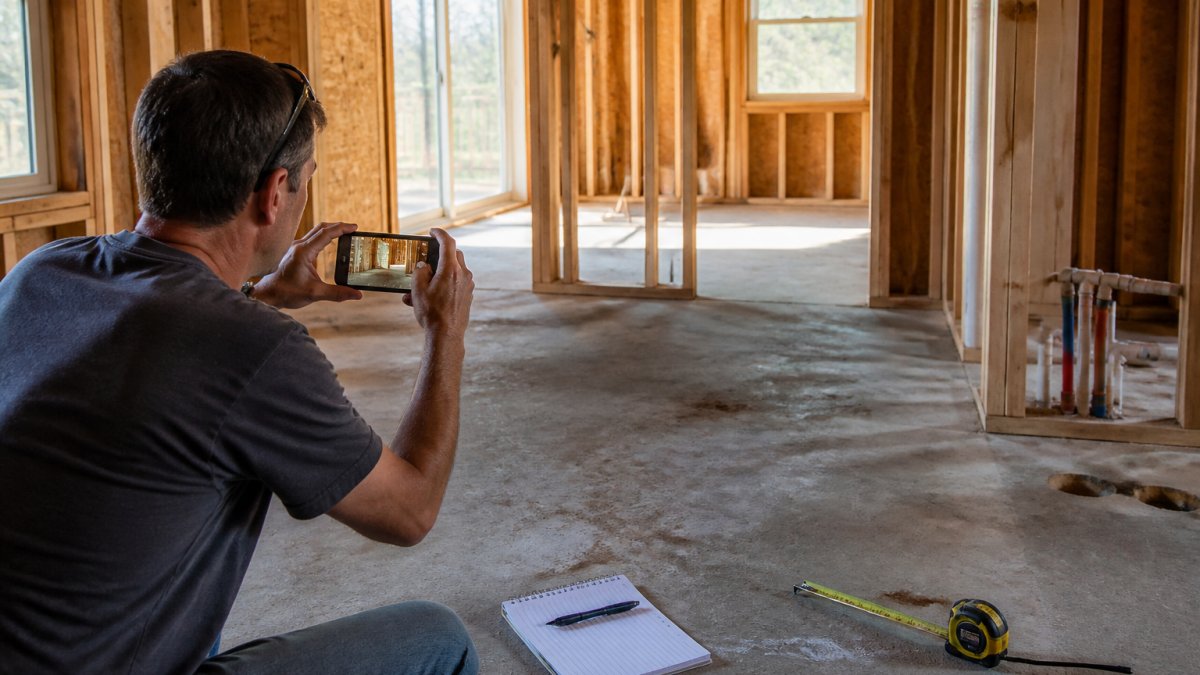 Homeowner photographing incomplete construction work on a job site with a notepad and smartphone visible, documenting unfinished tasks and measuring tape on floor