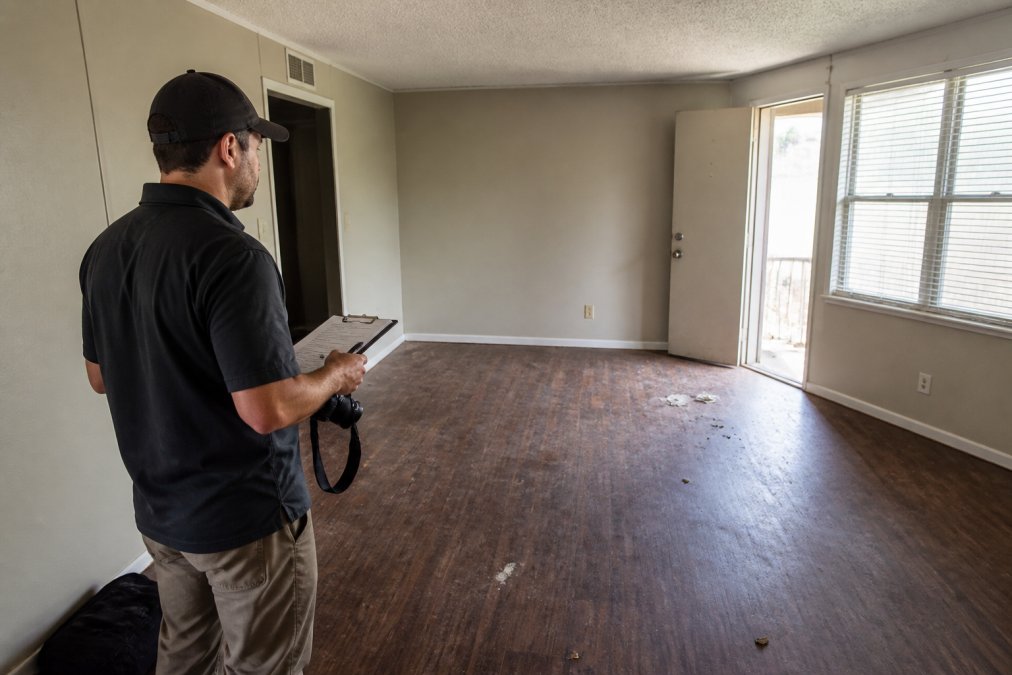 Property manager photographing an empty apartment room with a clipboard and camera, documenting vacant unit condition with natural light from windows