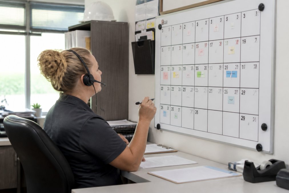 An HVAC dispatcher wearing a headset reviewing a large wall calendar filled with appointments in a bright, organized dispatch office during summer
