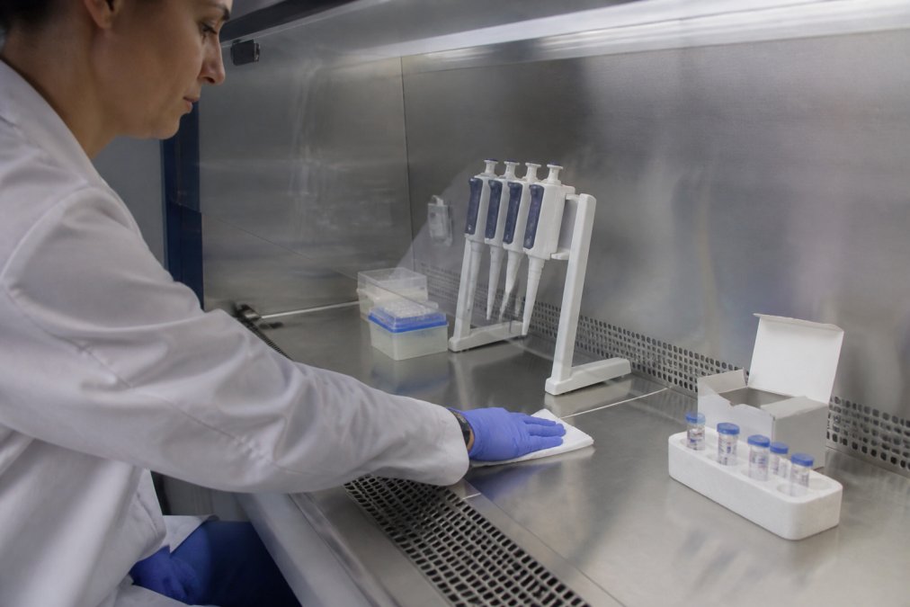 A researcher in a white lab coat and nitrile gloves carefully wiping down the stainless steel surface of a laminar flow hood with isopropyl alcohol wipes, sterile pipettes and sealed peptide vials visible on the workspace