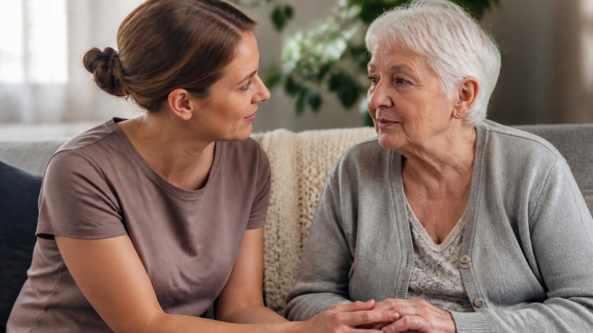 A carer sitting beside an older person in a softly lit, quiet room with warm natural lighting, maintaining calm eye contact and speaking gently with a reassuring, patient expression