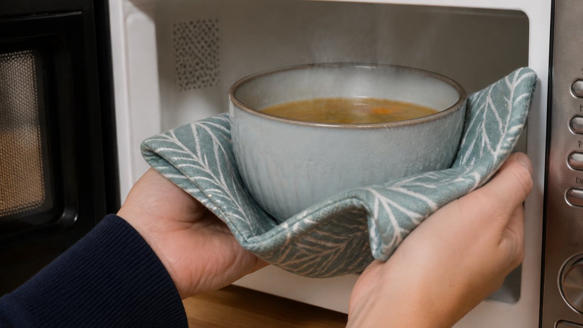 Close-up of hands holding a fabric bowl cozy while removing a steaming ceramic bowl from a microwave, showing the cozy in practical use with condensation visible on the bowl