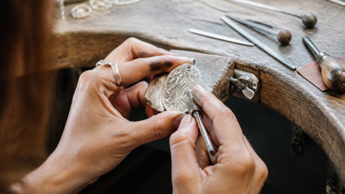 Close-up of a silversmith's hands shaping a silver pendant with jeweler's tools on a wooden workbench, natural studio lighting illuminating the intricate metalwork
