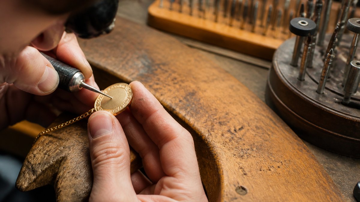Close-up of a jewelry artisan's hands using a fine engraving tool on a delicate gold necklace pendant, with a workbench of small tools and soft studio lighting in the background