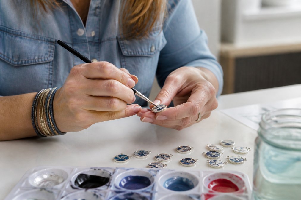 Close-up of a woman's hands carefully hand-painting a small sterling silver charm at a well-lit craft table, with several finished artisan-made charms arranged nearby on a white surface