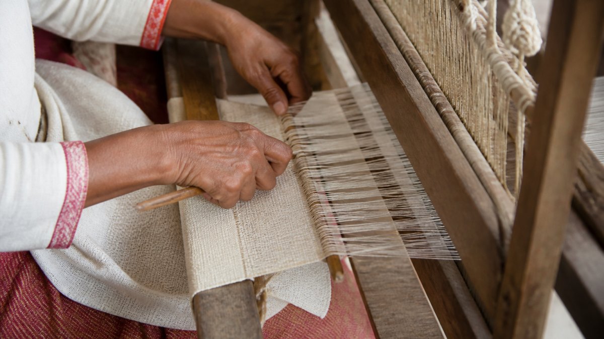 Close-up of hands working on traditional wooden loom with natural fiber threads, showing detailed weaving technique and textile construction in natural studio lighting
